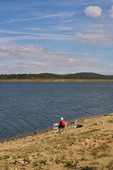 Fototapeta na wymiar Caucasian senior woman sitting on a chair with a straw hat on a dam lake reservoir in Alentejo, Portugal