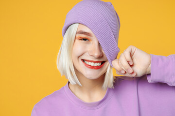 Smiling young funny blonde caucasian woman 20s bob haircut bright makeup wearing basic casual purple shirt covering eye with violet beanie hat isolated on yellow color background studio portrait.