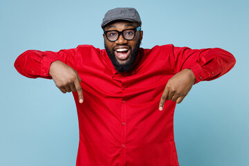 Surprised cheerful young bearded african american man 20s in casual red shirt eyeglasses cap pointing index fingers down on mock up copy space isolated on pastel blue color background studio portrait. © ViDi Studio