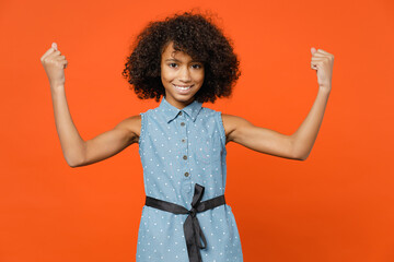 Smiling strong little african american kid girl 12-13 years old in denim dress showing biceps muscles isolated on bright orange color background children studio portrait. Childhood lifestyle concept.