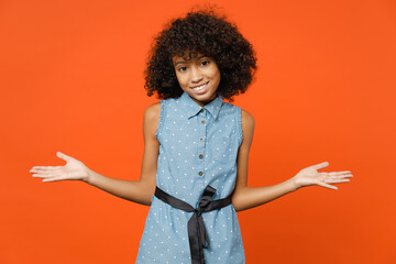 Confused smirked little african american kid girl 12-13 years old wearing casual denim dress spreading hands isolated on orange color background children studio portrait. Childhood lifestyle concept.