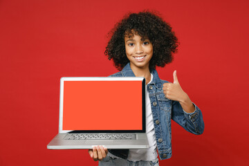 Naklejka na ściany i meble Funny little african american kid girl 12-13 in denim jacket hold laptop pc computer with blank empty screen showing thumb up isolated on red background children portrait. Childhood lifestyle concept.