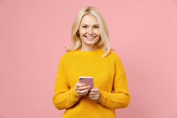 Smiling elderly gray-haired blonde woman lady 40s 50s years old in yellow casual sweater standing using mobile cell phone typing sms message isolated on pastel pink color background studio portrait.