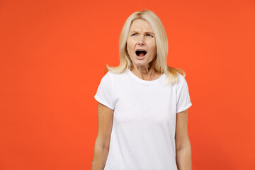 Shocked amazed elderly gray-haired blonde woman lady 40s 50s years old in white casual t-shirt standing keeping mouth open looking aside isolated on bright orange color background studio portrait.