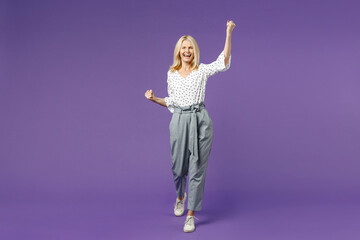 Full length of happy joyful elderly gray-haired blonde woman lady 40s 50s in white dotted blouse doing winner gesture clenching fists looking camera isolated on violet background studio portrait.