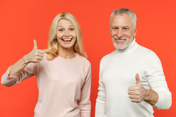 Excited cheerful funny couple two friends elderly gray-haired man blonde woman wearing white pink casual clothes standing showing thumbs up isolated on bright orange color background studio portrait.