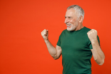 Smiling joyful elderly gray-haired mustache bearded man wearing basic casual green t-shirt standing doing winner gesture clenching fists isolated on bright orange color background studio portrait.