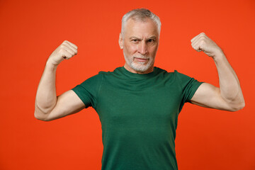Strong elderly gray-haired mustache bearded man wearing basic casual green t-shirt standing showing biceps muscles looking camera isolated on bright orange color wall background studio portrait.