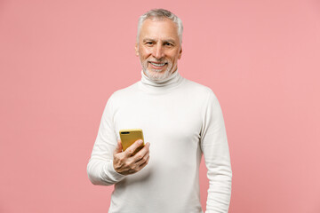 Smiling elderly gray-haired mustache bearded man in casual white turtleneck standing using mobile cell phone typing sms message looking camera isolated on pastel pink color background studio portrait.