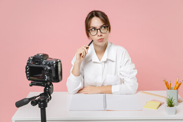 Pensive young woman tutor teacher in shirt glasses sit at desk looking aside conducting online lesson seminar recording video on camera isolated on pink background. Distance remote education concept.