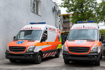 Two modern white and orange ambulance medical emergency vans truck parked near hospital building. Paramedic vehicles on city street. Healthcare and urgent medical support concept