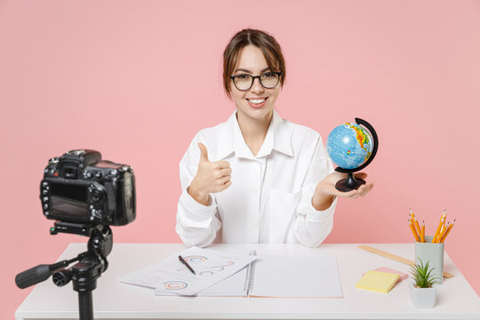 Smiling woman tutor teacher sit desk hold Earth world globe showing thumb up conducting online lesson seminar recording video on camera isolated on pink background. Distance remote education concept.