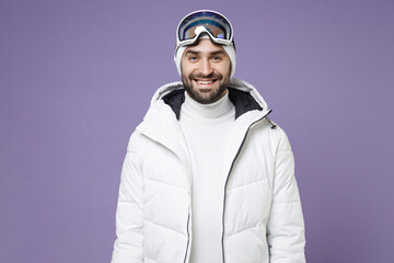 Smiling skier man in warm white padded windbreaker jacket ski goggles mask going to spend extreme weekend winter season in mountains isolated on purple background. People lifestyle hobby concept.