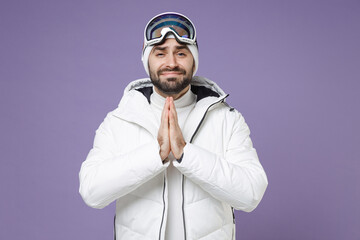 Pleading skier man in white windbreaker jacket ski goggles mask hold hands folded in prayer spend extreme weekend winter in mountains isolated on purple background. People lifestyle hobby concept.