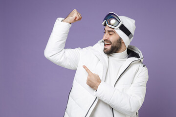 Funny skier man in white windbreaker jacket ski goggles mask pointing on biceps muscles spend extreme weekend winter season in mountains isolated on purple background. People lifestyle hobby concept.