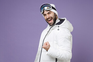Joyful skier man in white windbreaker jacket ski goggles mask clenching fist like winner spend extreme weekend winter season in mountains isolated on purple background. People lifestyle hobby concept.