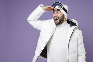 Amazed skier man in windbreaker jacket ski goggles mask hold hand at forehead looking far away distance spend extreme weekend winter in mountains isolated on purple background. People hobby concept.