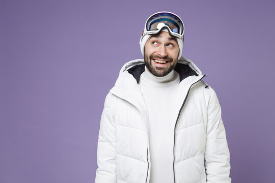 Pensive skier man in warm white windbreaker jacket ski goggles mask looking up going to spend extreme weekend winter season in mountains isolated on purple background. People lifestyle hobby concept.
