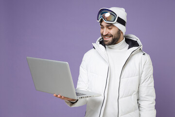 Smiling skier man in warm white windbreaker jacket ski goggles mask work on laptop pc computer spend extreme weekend winter in mountains isolated on purple background. People lifestyle hobby concept.