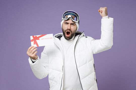 Happy skier man in white windbreaker jacket ski goggles mask hold gift certificate doing winner gesture spend weekend winter in mountains isolated on purple background. People lifestyle hobby concept.