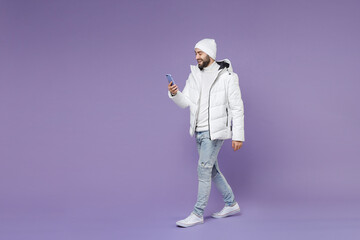 Full length side view smiling man in warm white windbreaker jacket hat using mobile cell phone typing sms message isolated on purple background studio portrait. People lifestyle cold season concept.