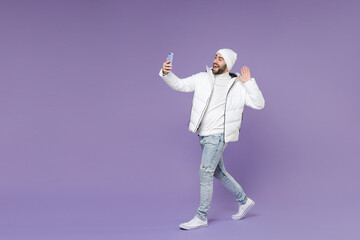 Full length side view of excited man in warm white windbreaker jacket hat doing selfie shot on mobile phone talk making video call isolated on purple background. People lifestyle cold season concept.