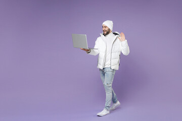 Full length funny man in warm white padded windbreaker jacket hat work on laptop pc computer making video call greeting with hand isolated on purple background. People lifestyle cold season concept. © ViDi Studio