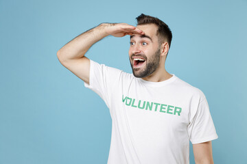 Excited young man in white volunteer t-shirt holding hand at forehead looking far away distance...
