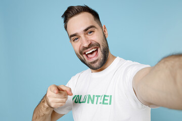 Close up excited young bearded man in volunteer t-shirt doing selfie shot on mobile phone point finger on camera isolated on blue background. Voluntary free work assistance help charity grace concept.