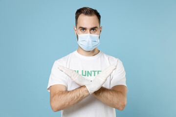 Man in volunteer t-shirt gloves face mask safe from coronavirus virus covid-19 hold crossed hands in stop gesture isolated on blue background. Voluntary free work assistance aid help support concept.