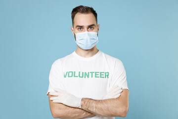Young man in volunteer t-shirt sterile gloves face mask to safe from coronavirus virus covid-19 hold hands crossed isolated on blue background. Voluntary free work assistance aid help support concept.