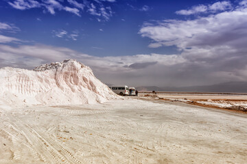 Mountain of pink salt with traces of an excavator. Two minibuses are parked near the mountain and people are standing nearby. Mountains on the horizon with a background of blue sky with clouds.