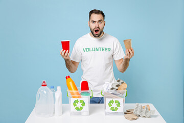 Shocked worried young man in volunteer t-shirt near recycling stations sorting plastic paper trash hold cups keep mouth open isolated on blue background. Voluntary free work assistance help concept.