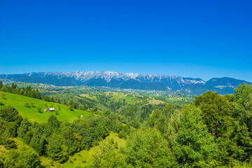 Fototapeta na wymiar Stunning alpine landscape with green fields and high snowy Piatra Craiului mountains near Brasov. Mountain farm with old wooden house. Bran, Transylvania, Romania, Europe. 