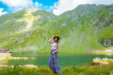 A beautiful woman in a long dress holding straw hat on the shores of Lake Balea against the backdrop of the high Romanian mountains. exciting holidays in the mountains in Transylvania, Romania