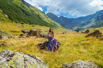 Fototapeta premium Sensual pretty girl in long dress sits on a rock against the high mountains backgdrop. Woman sitting near Transfegerasan, one of the most spectacular mountain road in the world. Romanian summer travel