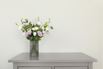 Bouquet of beautiful Eustoma flowers on chest of drawers near light wall. Space for text