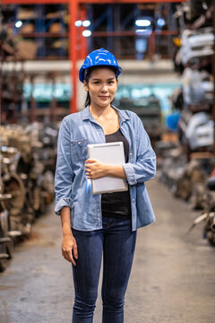 Confident engineer woman workers wear a safety helmet and standing in the automotive spare parts warehouse. Holding a tablet and smile. Check stock and quality concept. Mamy old engine in site