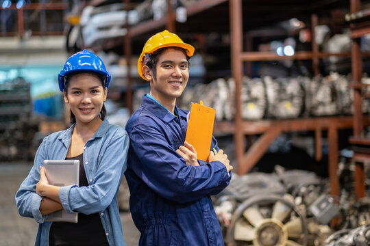 Engineer workers wear a safety helmet and crossed arms standing in the automotive spare parts warehouse. Maintainance and service concept. Man holding clipboard and women holding the tablet.