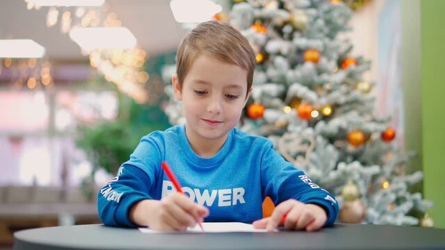 The boy writes a letter to Santa Claus and smiles against the background of a Christmas tree in flashing decorations. High quality FullHD footage