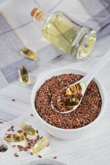 Brown flax seed or linseed in small bowl and gelatin capsules with omega oil on a white wooden background.