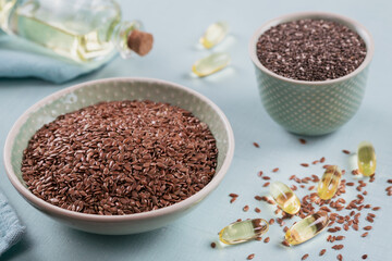 Brown flax seed or linseed and chia in small bowl and gelatin capsules with omega oil on a light blue background.