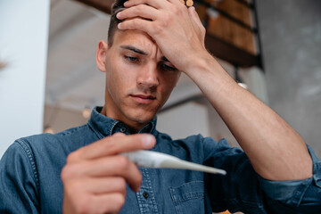 young man with a headache looking at the digital thermometer reading.