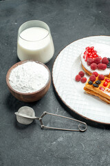 bottom view tart and waffles with berries on white plate a bowl of powdered sugar a sieve and a glass of milk on dark background
