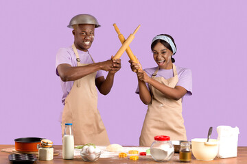 Silly African American couple having mock fight with rolling pins while baking in kitchen, lilac...