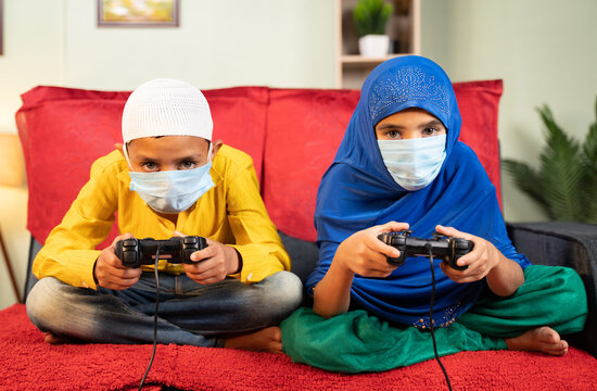 Two Muslim Kids With Medical Face Mask Busy Playing Video Game Using Gamepad At Home - Concept Of Kids On Game During Coronavirus Covid-19 Lockdown.