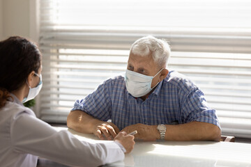 Mature 80s male patient in medical facemask have consultation with female doctor in hospital. Woman GP or therapist talk consult with senior man client in facial mask. Elderly healthcare concept.