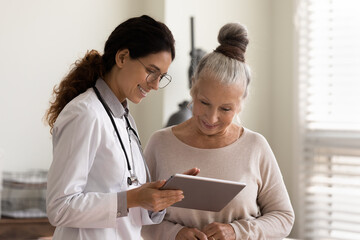 Happy female doctor and senior patient look at tablet screen discuss treatment or therapy on...