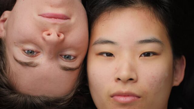 Young Asian and Caucasian woman waking up, opening eyes and looking at camera. Two girls lying down side-by-side. Concept of interracial friendship, female mixed race homosexual couple. Close up, 4K.