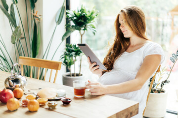 A pregnant woman is eating breakfast at the table and looking into a tablet. Communication and work on the Internet.